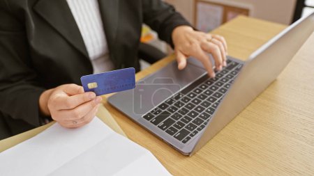 Foto de Una mujer de negocios en una chaqueta utiliza una tarjeta de crédito en un ordenador portátil para una transacción en línea en una oficina. - Imagen libre de derechos