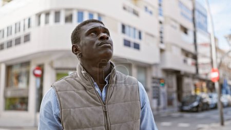 Foto de Un hombre africano contemplativo de pie en una calle de la ciudad con edificios y coches en el fondo - Imagen libre de derechos