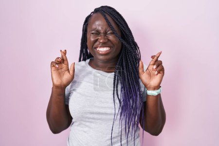 Téléchargez les photos : Jeune femme africaine debout sur fond rose geste doigt croisé souriant avec espoir et les yeux fermés. chance et concept superstitieux. - en image libre de droit