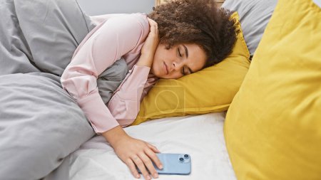 Foto de Una joven hispana con el pelo rizado duerme tranquilamente en un dormitorio, con la mano apoyada en un smartphone. - Imagen libre de derechos