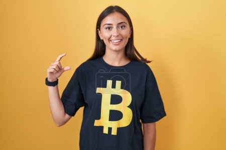 Photo for Young hispanic woman wearing bitcoin t shirt smiling and confident gesturing with hand doing small size sign with fingers looking and the camera. measure concept. - Royalty Free Image