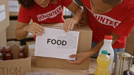 Photo for Young and beautiful man and woman volunteers united in altruism, working together, putting paper wrapped food into package for charity donations at community center - Royalty Free Image