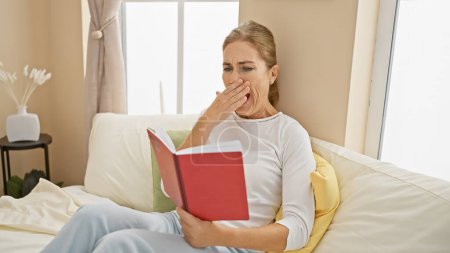 Photo for Caucasian woman yawns while reading a red book, sitting on a sofa in a cozy, well-lit home interior - Royalty Free Image