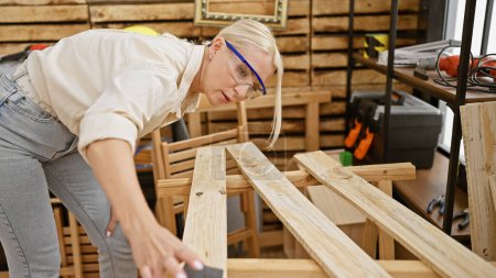 Foto de Joven mujer rubia carpintero con gafas de seguridad lijado tablón de madera en la carpintería - Imagen libre de derechos