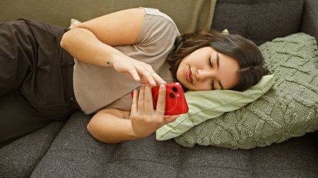Téléchargez les photos : Une femme hispanique détendue se prélasse à la maison, utilisant son smartphone sur le canapé, incarnant loisirs et style de vie moderne. - en image libre de droit