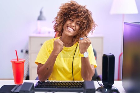 Téléchargez les photos : Jeune femme hispanique aux cheveux bouclés jouant à des jeux vidéo portant des écouteurs très heureux et excité de faire geste gagnant avec les bras levés, souriant et criant pour le succès. concept de célébration. - en image libre de droit