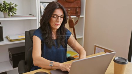 Téléchargez les photos : Une femme latina professionnelle d'âge moyen se concentre sur son ordinateur portable dans un cadre de bureau moderne. - en image libre de droit
