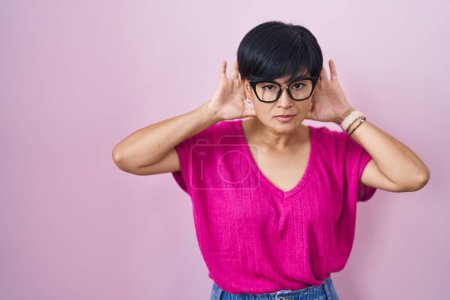Téléchargez les photos : Jeune femme asiatique aux cheveux courts debout sur fond rose essayant d'entendre les deux mains sur le geste de l'oreille, curieuse de commérages. problème auditif, sourd - en image libre de droit