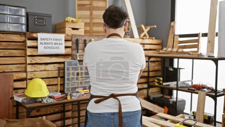 Foto de Vista posterior de un hombre con un delantal en un taller de carpintería rodeado de herramientas y madera. - Imagen libre de derechos