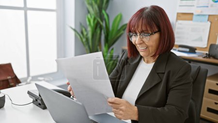 Téléchargez les photos : Une femme hispanique d'âge moyen joyeuse examine un document dans un cadre de bureau lumineux, transmettant professionnalisme et engagement. - en image libre de droit