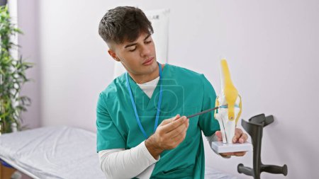 Foto de Hombre hispano joven guapo - un fisio inteligente en la clínica de rehabilitación, explicando profundamente la estructura y el tratamiento de la rodilla usando un modelo anatómico - Imagen libre de derechos