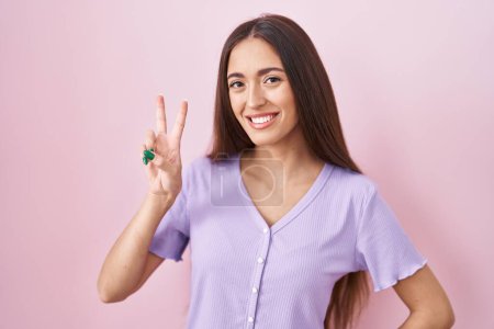 Téléchargez les photos : Jeune femme hispanique aux cheveux longs debout sur fond rose souriant en regardant la caméra montrant des doigts faisant signe de victoire. numéro deux. - en image libre de droit