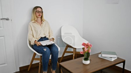 Foto de Joven mujer rubia trabajadora de negocios sosteniendo portapapeles sentada en silla con cara seria en la sala de espera de la oficina - Imagen libre de derechos