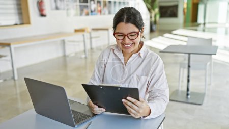 Photo for A cheerful young hispanic woman working with a laptop and clipboard in a modern office interior. - Royalty Free Image