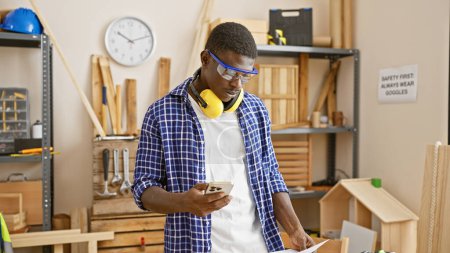 A focused african man in protective gear using a smartphone in a carpentry workshop.