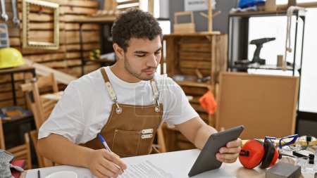 Photo for A thoughtful man in a woodworking studio examines a tablet beside safety gear and papers. - Royalty Free Image