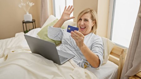 Téléchargez les photos : Femme d'âge moyen souriante avec carte de crédit agitant à l'ordinateur portable dans la chambre, représentant des achats en ligne ou salutation virtuelle. - en image libre de droit