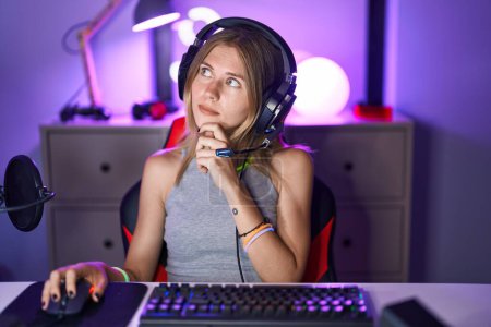 Photo for Blonde caucasian woman playing video games with headphones serious face thinking about question with hand on chin, thoughtful about confusing idea - Royalty Free Image