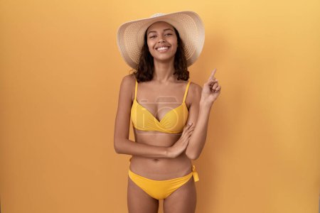 Photo for Young hispanic woman wearing bikini and summer hat with a big smile on face, pointing with hand finger to the side looking at the camera. - Royalty Free Image