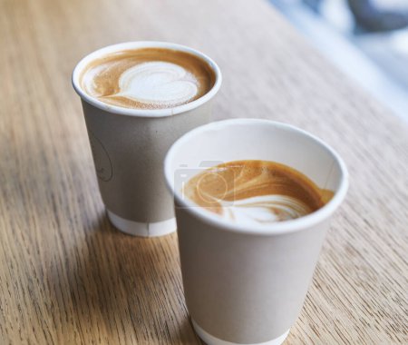 Téléchargez les photos : Deux tasses à emporter en papier de cappuccino avec latte art en forme de cœur assis sur une table en bois. - en image libre de droit