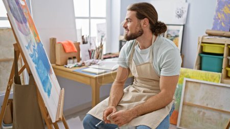 Téléchargez les photos : Jeune homme hispanique artiste souriant confiant assis sur la chaise au studio d'art - en image libre de droit