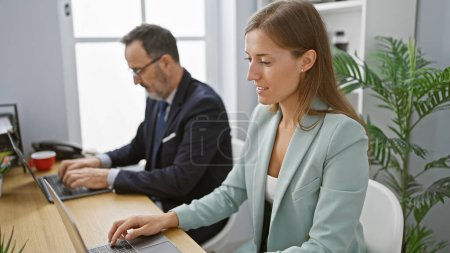 Foto de Dos trabajadores de negocios sonrientes sentados juntos en el escritorio de la oficina, trabajando con confianza en la computadora portátil con su atuendo profesional - Imagen libre de derechos