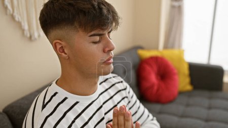 Téléchargez les photos : Jeune homme hispanique attrayant trouve l'équilibre dans la foi, priant les yeux fermés sur son canapé du salon, un souffle de calme et de liberté remplit sa maison comme le portrait d'un gars détendu méditant à l'intérieur - en image libre de droit