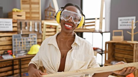 Foto de Hermosa mujer afroamericana carpintera, adornada con gafas de seguridad y auriculares, manejando magistralmente tablón de madera en el bullicioso taller de carpintería - Imagen libre de derechos