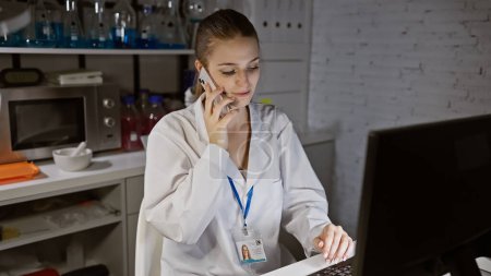 A young woman in a lab coat is engaged in a phone conversation while using a laptop in a laboratory setting.