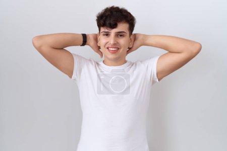 Téléchargez les photos : Jeune homme non binaire portant un t-shirt blanc décontracté relaxant et étirant, bras et mains derrière la tête et le cou souriant heureux - en image libre de droit