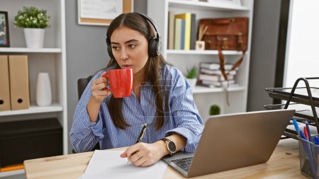 Foto de Una mujer hispana disfrutando del café mientras trabaja remotamente desde una oficina bien organizada. - Imagen libre de derechos