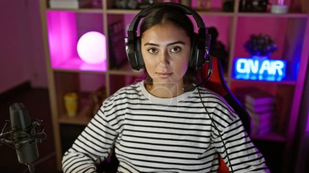 Photo for Hispanic woman with headphones in a gaming room illuminated by neon lights at night, projecting a contemporary vibe. - Royalty Free Image