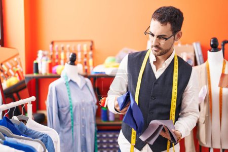Young hispanic man tailor holding cloths at atelier