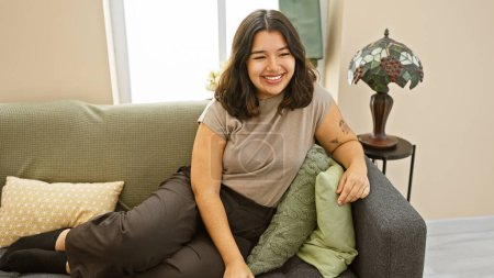 Téléchargez les photos : Jeune femme hispanique souriante se relaxant sur un canapé dans un cadre confortable de salon, rayonnant de beauté et de contentement. - en image libre de droit