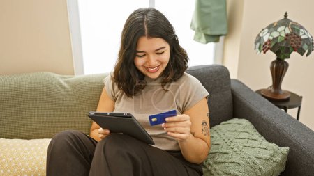 Foto de Mujer hispana sonriendo mientras hace compras en línea con tableta y tarjeta de crédito en una acogedora sala de estar. - Imagen libre de derechos