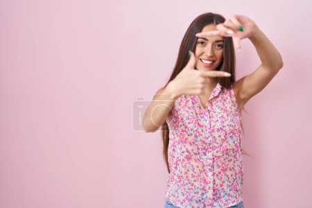 Téléchargez les photos : Jeune femme hispanique aux cheveux longs debout sur fond rose souriant faisant cadre avec les mains et les doigts avec le visage heureux. concept de créativité et photographie. - en image libre de droit