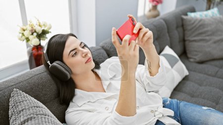 Eine attraktive junge hispanische Frau lümmelt mit Kopfhörern und benutzt lässig ein Smartphone in einer gemütlichen Wohnzimmeratmosphäre.