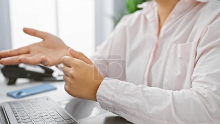 Mujer latina joven en un entorno de oficina que experimenta dolor de muñeca posiblemente debido a la tensión repetitiva del uso de la computadora.