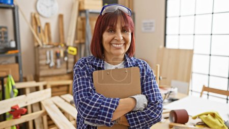 Foto de Mujer hispana madura sonriendo con confianza en un taller de carpintería ordenado, sosteniendo un portapapeles. - Imagen libre de derechos