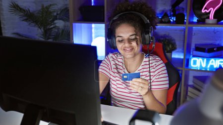 Photo for A young hispanic woman with curly hair smiling at a credit card while gaming indoors at night. - Royalty Free Image