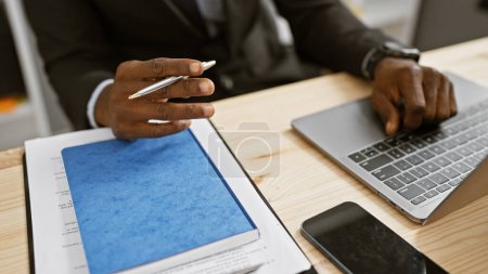Photo for African american businessman working on laptop with pen and folder in modern office setting. - Royalty Free Image