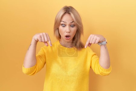 Foto de Mujer joven caucásica con suéter amarillo apuntando hacia abajo con los dedos mostrando publicidad, cara sorprendida y boca abierta - Imagen libre de derechos