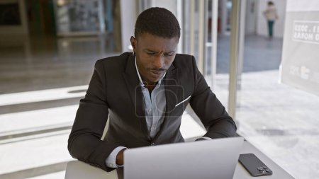 Foto de Un hombre afroamericano enfocado en un traje que trabaja en una computadora portátil en un ambiente de oficina moderno, transmitiendo profesionalismo y dedicación. - Imagen libre de derechos
