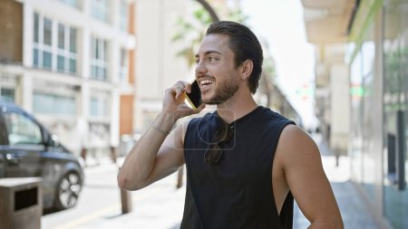 Téléchargez les photos : Jeune homme hispanique parlant sur smartphone souriant à la rue - en image libre de droit