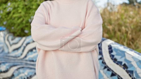 Téléchargez les photos : Femme confiante en pull rose se tient avec les bras croisés dans un parc urbain décoré de graffitis. - en image libre de droit
