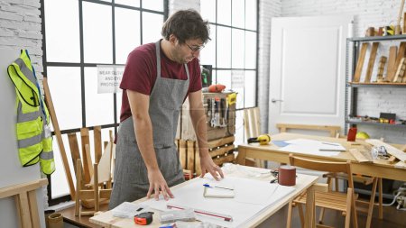 Foto de Un hombre hispano enfocado revisa planes en un taller de carpintería soleado, representando artesanía y habilidad. - Imagen libre de derechos