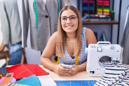 Téléchargez les photos : Jeune belle hispanique femme tailleur souriant confiant en utilisant smartphone au magasin de tailleur - en image libre de droit