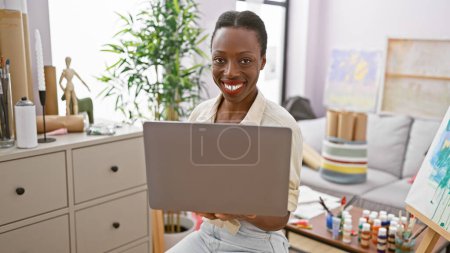 Photo for Confident beautiful african american woman artist, drawing inspiration via laptop, with a captivating smile lighting up the art studio - Royalty Free Image