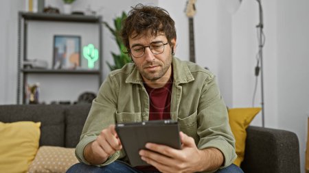 A focused man with glasses and a beard uses a tablet while sitting on a couch at home, hinting at leisure or remote work.