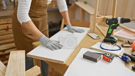 Téléchargez les photos : Une femme dans un atelier manipulant du bois avec une perceuse et des gants de sécurité à proximité, indiquant un travail de menuiserie. - en image libre de droit
